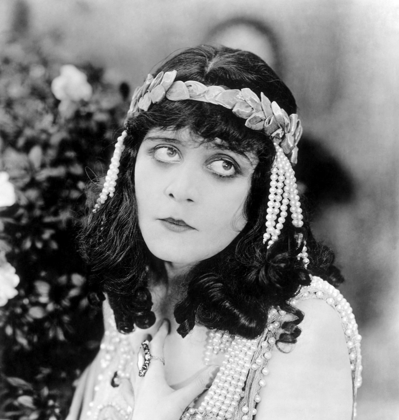 1918: American actor Theda Bara (1885-1955) looks up, holding one hand to her chest in a still from director J. Gordon Edwards' silent film 'Salome'. She wears a garland headband with pearls hanging from her hair and shoulders.